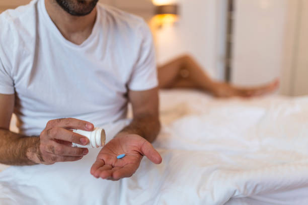 Mens health and sexual problems, male power and libido. Focus on blue pill in hands of happy young man, blurred smiling wife sits in bed in modern interior of bedroom, close up stock photo
