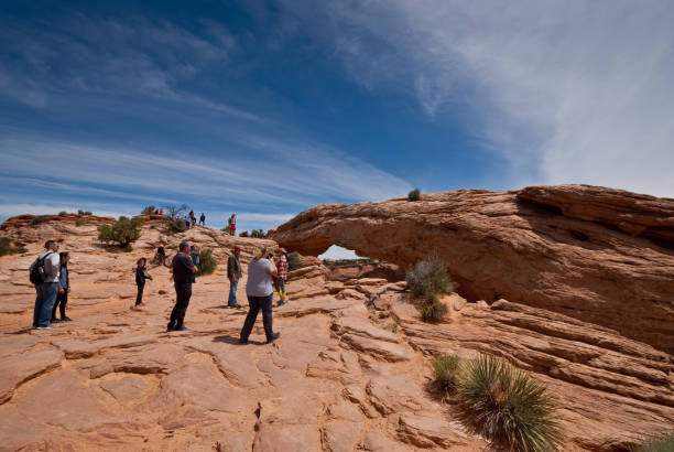 People at Mesa Arch Canyonlands National Park, Utah, USA - May 29, 2019: Mesa Arch is a pothole arch, formed when rainwater eventually broke the bonds between grains of sand and deepened surface depressions into a hole. This arch is also geologically unusual in that it sits at the top of a canyon wall. Eventually the same forces of water freezing and thawing will lead to the collapse of this arch. In this picture people are lining up to take their picture with the famous arch. Mesa Arch is on the eastern edge of the Island in the Sky Mesa. jeff goulden canyonlands national park stock pictures, royalty-free photos & images