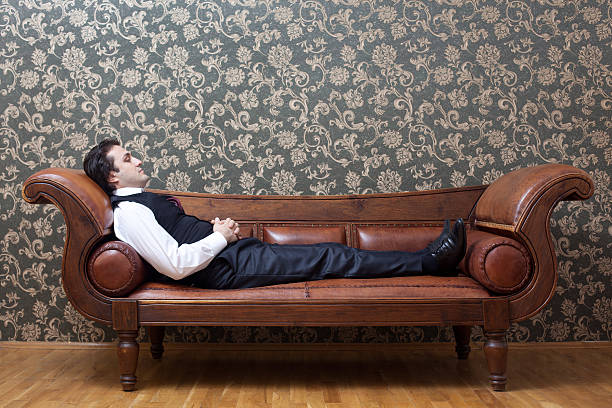 Mesmerized man lying down on coach in psychiatrist office A young mesmerized man is lying down on brown leather coach in  psychiatrist office.The wall is wallpapered.The model and coach is seen in full length.He is in formal clothes and his hands are on his belly and his eyes are closed.The image was shot with a full frame DSLR camera. therapist couch stock pictures, royalty-free photos & images