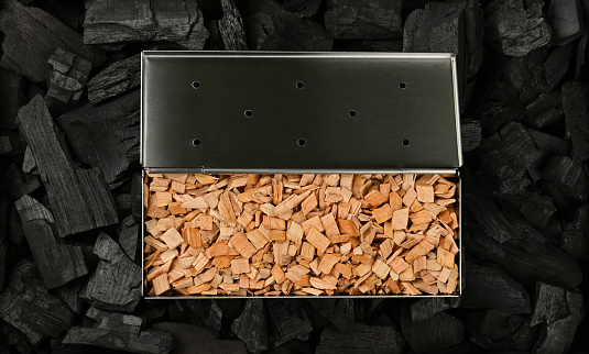 Close up one stainless steel metal smoker box with hardwood alder chips on black lump charcoal pieces, ready to barbecue grill and smoke food, elevated top view, directly above