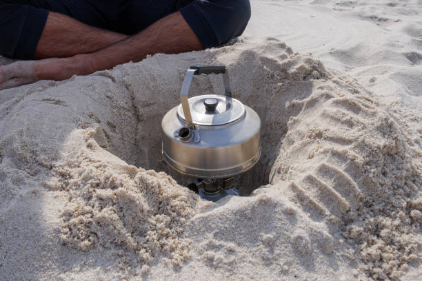 Metal teapot on camping kettle fire burner in a hole dug in the sand on a beach. Travel camping outdoor portable hot water. stock photo