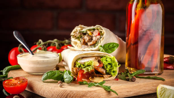 Mexican cuisine, burrito with chicken, cherry tomatoes, lettuce, mushrooms, rucola and chili peppers, on a black background. With salsa sauce. Copy space, selective focus stock photo