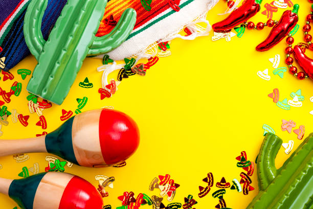 Mexican fiesta and Cinco de Mayo party concept theme with jalapeno pepper necklace, maracas, cactus and traditional rug covered in sombrero shaped confetti on yellow background with copy space stock photo