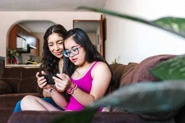 Mexican girl with down syndrome and her mother spending time together using the phone at home, in disability concept in Latin America stock photo