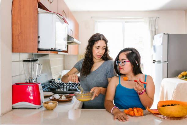 Mexican teen girl with down syndrome eating fruit and her mother cooking at home, in disability concept in Latin America stock photo