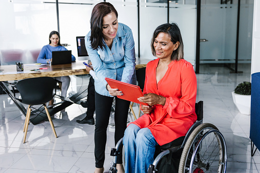 Mexican transgender woman in a wheelchair with young office colleague sharing information at the workplace in Latin America