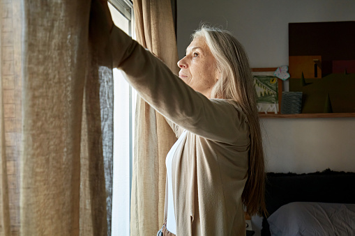 Partial side view of woman with long gray hair opening curtain at bedroom window and looking at view.