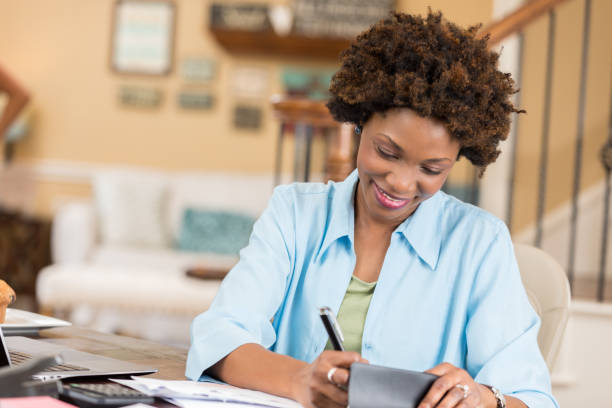 Mid adult African American woman balances checkbook in home office Pretty African American woman concentrates while writing a check or balancing her checkbook. Personal bills or documents are on the desk along with a laptop. bank account stock pictures, royalty-free photos & images