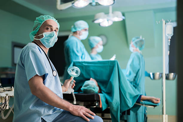Mid adult anesthesiologist during surgery in operating room. Male doctor sitting in operating room during surgery while holding oxygen mask and looking at the camera. anesthetic stock pictures, royalty-free photos & images
