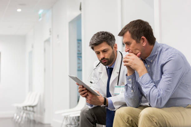 Mid adult doctor explaining serious patient over digital tablet at hospital stock photo