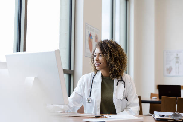 mid adult female doctor reviews patient records on desktop pc - doutor imagens e fotografias de stock