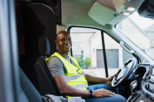 The mid adult gig worker smiles from the driver's seat of his delivery van.
