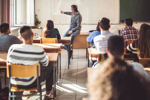 Mid adult male teacher giving a lecture to his students in the classroom. Male professor sitting on the table and teaching mathematics from whiteboard at high school. high school building stock pictures, royalty-free photos & images