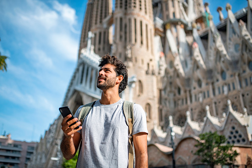 Low angle view of 30 year old Hispanic male tourist using smart phone for sightseeing guidance with Sagrada Familia in background.