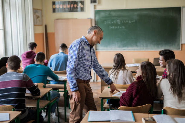 Mid adult professor helping his students during a class at high school. Happy male teacher assisting teenage students in the classroom. high school building stock pictures, royalty-free photos & images