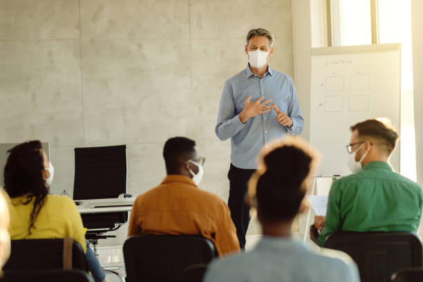 Mid adult teacher with protective face masks talking to a group of university students in lecture hall. University professor wearing protective face mask while holding a class to group of students during coronavirus epidemic. professor stock pictures, royalty-free photos & images
