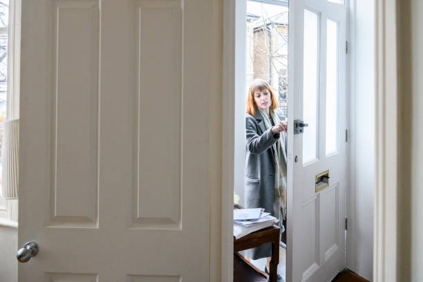Mid adult woman unlocking front door and arriving home Businesswoman coming home at the end of the working day, view from inside house with internal door in foreground, woman seen through gap in front door worker returning home stock pictures, royalty-free photos & images