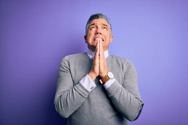 Middle age handsome grey-haired man wearing elegant sweater over purple background begging and praying with hands together with hope expression on face very emotional and worried. Begging.  prayer request stock pictures, royalty-free photos & images