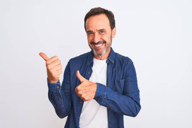 middle age handsome man wearing blue denim shirt standing over isolated white background pointing to the back behind with hand and thumbs up, smiling confident - man pointing imagens e fotografias de stock