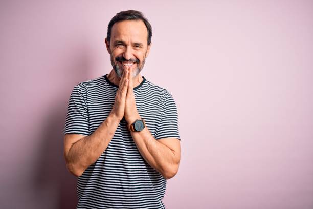 Middle age hoary man wearing casual striped t-shirt standing over isolated pink background praying with hands together asking for forgiveness smiling confident.  prayer request stock pictures, royalty-free photos & images