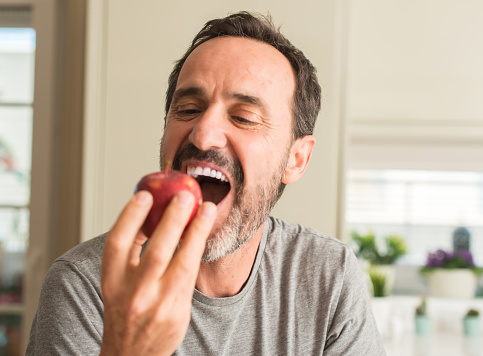 Middle age man eating healthy red apple with a happy face standing and smiling with a confident smile showing teeth