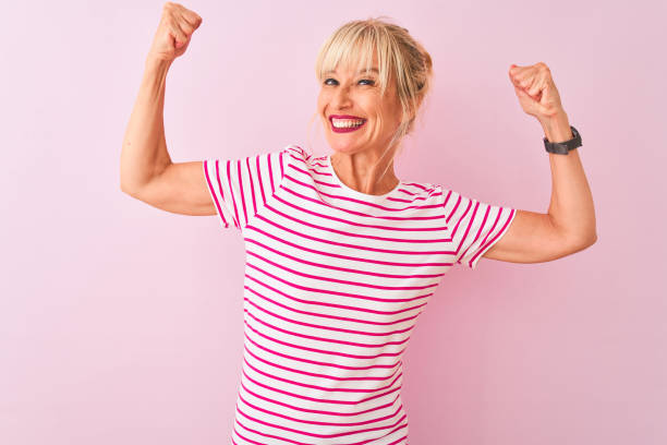 mujer de mediana edad con camiseta a rayas de pie sobre fondo rosa aislado que muestra los músculos de los brazos sonriendo orgulloso. concepto de acondicionamiento físico. - atlético fotografías e imágenes de stock