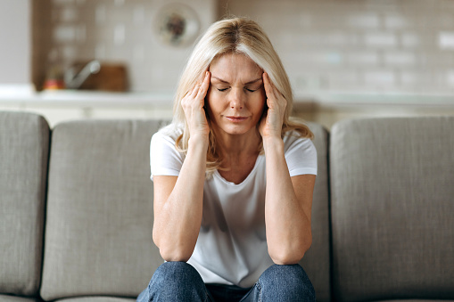 Middle aged blonde woman sits on couch at living room holding her head with her hands, feels unhappy because of headache, personal troubles, illness or bad news, she need psychological or medical support