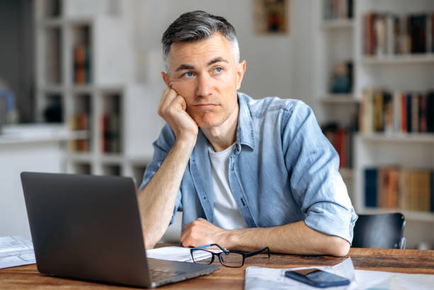 Middle aged caucasian business man or freelancer sitting at workplace, working on laptop, resting head on hand because of tired, doing work overtime, stressed and bored, need rest, pensive looks away stock photo