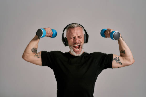 middle aged muscular man in black t shirt and headphones closing eyes, holding blue dumbbells, posing in studio over grey background - medial object imagens e fotografias de stock