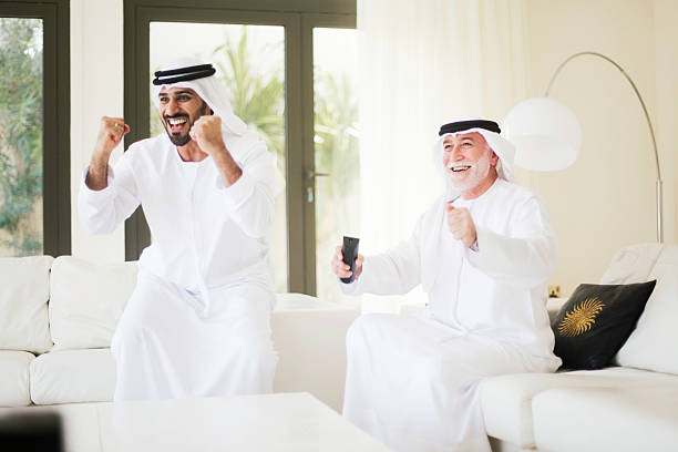 Middle eastern father and watching soccer on tv at home. Middle eastern senior watching tv at home dressed in dishdasha. old arab man stock pictures, royalty-free photos & images