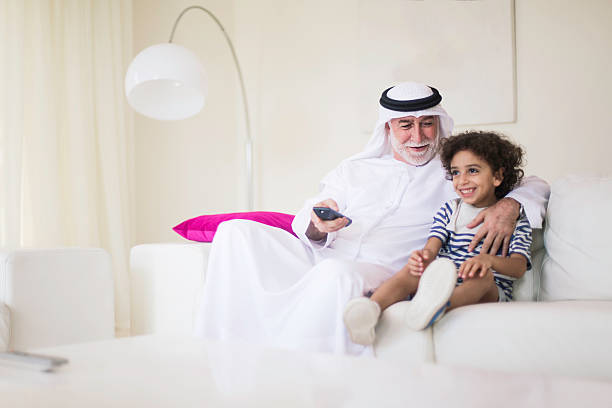 Middle eastern grandfather watching tv at home with grandson. Middle eastern senior watching tv at home dressed in dishdasha. old arab man stock pictures, royalty-free photos & images