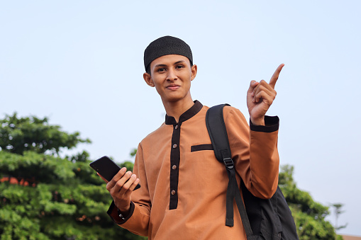 Religious smiling middle eastern man in brown muslim shirt and black cap pointing away with his finger while using mobile phone against park background