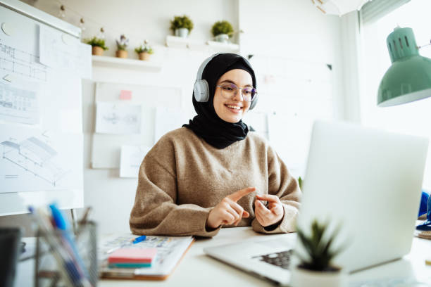 Middle Eastern woman working on laptop in office and having video call with partners Middle Eastern woman with black hijab working on laptop in office middle eastern woman stock pictures, royalty-free photos & images