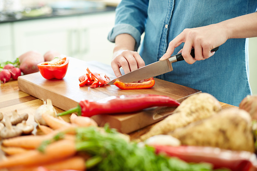 Midsection image of woman chopping red bell pepper on cutting board. There are various vegetables on wooden table. Focus is on her hands. Female is preparing food. She is in domestic kitchen.
