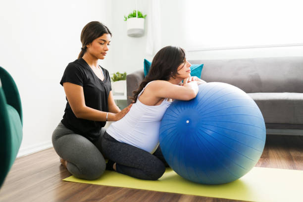 Midwife rubbing the back of a pregnant woman Beautiful doula in activewear helping a caucasian pregnant woman with her back pain. Expectant mother relaxing in a fitness ball while receiving a massage midwife stock pictures, royalty-free photos & images