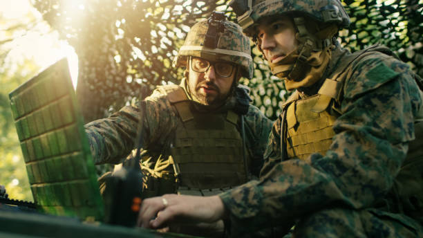 Military Staging Base, Officer Gives Orders to Chief Engineer, They Use Radio and Army Grade Laptop. They're in Camouflaged Tent in a Forest. They're on Reconnaissance Operation/ Mission. Military Staging Base, Officer Gives Orders to Chief Engineer, They Use Radio and Army Grade Laptop. They're in Camouflaged Tent in a Forest. They're on Reconnaissance Operation/ Mission. military base stock pictures, royalty-free photos & images