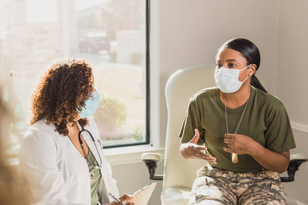 Military veteran discusses health with doctor A serious female army veteran gestures as she describes symptoms to a female doctor. They are wearing protective face masks amid the coronavirus pandemic. mental health professional photos stock pictures, royalty-free photos & images