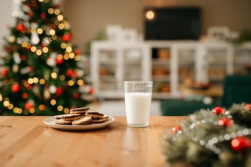 Still life photo of a plate with cookies and a glass of milk that is waiting for Santa Claus. In the back you can see a nice and cosy living room with decorated Christmas tree. No people. Copy space. Daylight.