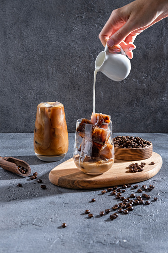 Milk Being Poured Into Iced Coffee on a dark table. Ice coffee in a tall glass with cream poured over and coffee beans. Cold coffee drink on a dark background with copy space. Set with different types of coffee drinks on a dark table.