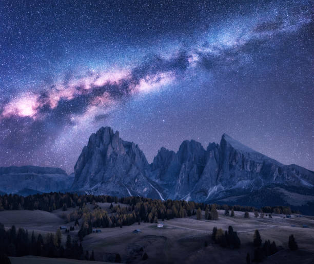 Photo of Milky Way over beautiful mauntains at night. Autumn landscape with mountains, purple sky with stars and bright milky way, buildings, trees, high rocks. Alpe di Siusi in Dolomites, Italy. Space