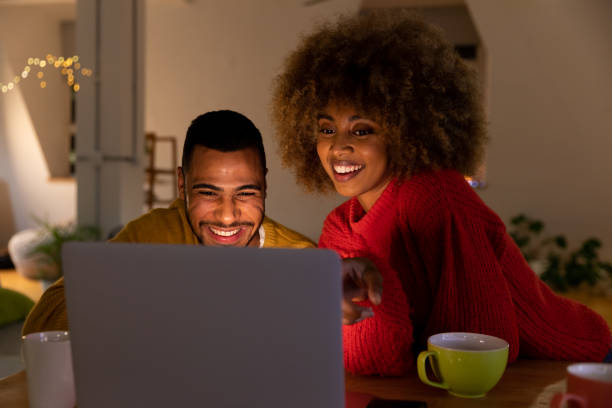 Millennial adult friends socialising together at home Front view close up of a young mixed race man and woman sitting at a table looking at a laptop computer together and smiling in the sitting room of an apartment in front of stock pictures, royalty-free photos & images