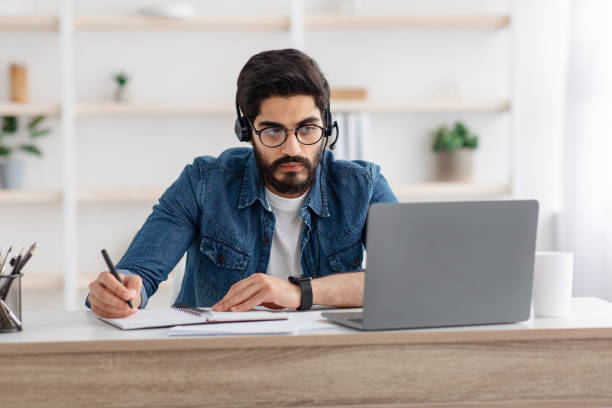 Millennial arab guy in headset watching online webinar on laptop at home, studying with computer and taking notes Millennial arab guy in headset watching online webinar on laptop at home, having online training, studying with computer and taking notes to notepad, copy space online computer science degree stock pictures, royalty-free photos & images