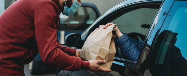 Millennial man is giving a lunch box to a woman in her car Millennial man is giving a lunch box to a woman in her car. View from outside the car. They are wearing protective face masks. curbsidepickup stock pictures, royalty-free photos & images