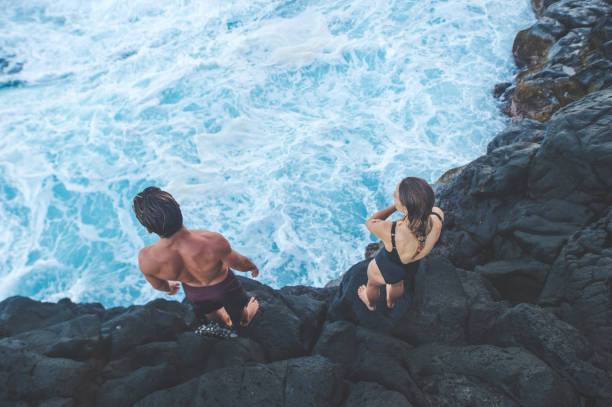 A millennial-age couple getting ready to jump into the ocean A millennial-age male and female stand at the edge of a cliff and get ready to leap into the ocean. The waves are crashing into the rocks below. The shot is from behind them. cliff jumping stock pictures, royalty-free photos & images
