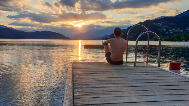 Millstaettersee - A man in full cup sitting at the end of a wooden pier of Millstaetter lake and enjoys the sunset. The sun sets A man in full cup sitting at the end of a wooden pier of Millstaetter lake and enjoys the sunset. The sun sets behind high Alps. Calm surface of the lake reflects the orange sky and the mountains alpine lakes wilderness stock pictures, royalty-free photos & images