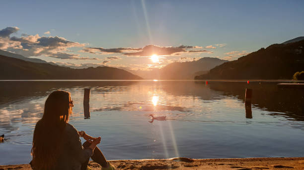 Millstaettersee - A woman in a jeans jacket sitting at the shore of Millstaettersee and enjoying the sunset. A few ducks swimming A woman in a jeans jacket sitting at the shore of Millstaettersee and enjoying the sunset. A few ducks swimming around. Calm surface of the lake reflects the orange sky and the mountains. High Alps. alpine lakes wilderness stock pictures, royalty-free photos & images