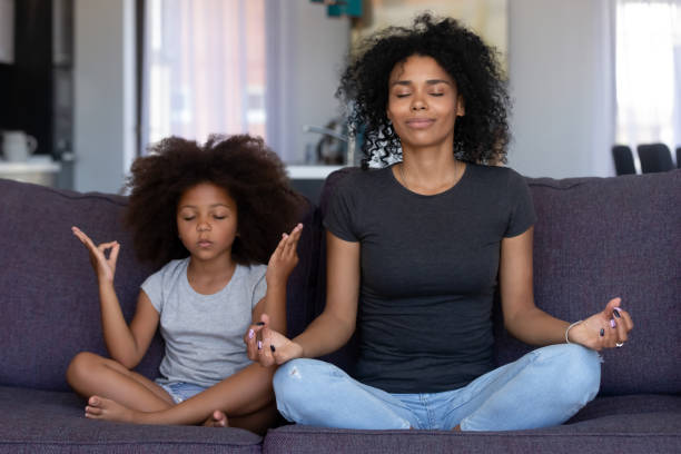 Mindful african mom with funny kid daughter doing yoga together Mindful african mom with cute funny kid daughter doing yoga exercise at home, calm black mother and mixed race little girl sitting in lotus pose on couch together, mum teaching child to meditate emotional stress photos stock pictures, royalty-free photos & images