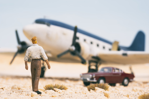 A miniature man in the desert eyes up a DC-3 Dakota aeroplane and a swish sports coupe next to it.