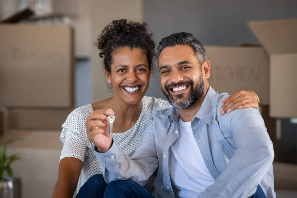 Mixed race couple holding new house keys Middle aged multiethnic couple embracing and holding house keys with carboard boxes behind them. Cheerful couple sitting on floor in new home. Portrait of indian man and african woman showing house keys while looking at camera after relocation. home ownership stock pictures, royalty-free photos & images