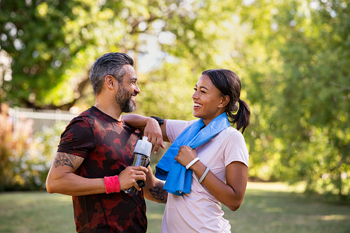 Cheerful multiethnic couple resting together after jogging in the park. Happy mature man and beautiful woman laughing while resting after running during summer afternoon. Smiling woman and carefree indian man taking a break after fitness training workout while looking at each other.